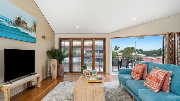 Amplia sala de estar con una gran televisión de pantalla plana, un sofá azul con cojines estampados, piso de madera y un amplio ventanal corredero que da a un balcón con vista al mar y a edificios.