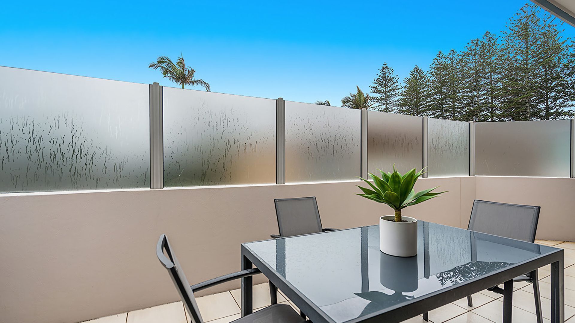 An outdoor patio with a glass table and chairs, a potted plant, and a frosted privacy wall.