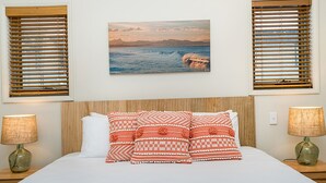 Un dormitorio con una cama, dos mesitas de noche, dos lámparas y un cuadro colgado en la pared que muestra una escena costera.