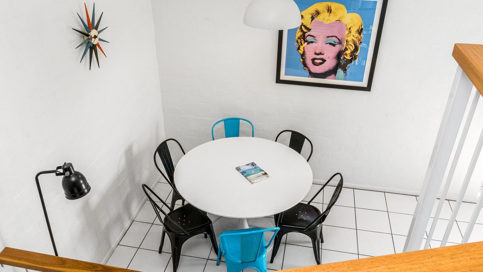 Un coin repas avec une table ronde, quatre chaises, une chaise bleue, une lampe noire et un tableau encadré au mur.