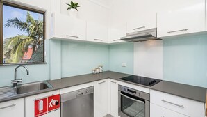 A modern kitchen with white cabinets, a stainless steel sink, a dishwasher, an oven, and a stove. There is a window with a view of palm trees and a red roof. - Byron Bay Accom - Balcony on Lawson - 6 - 34 Lawson Street (Byron Shire)