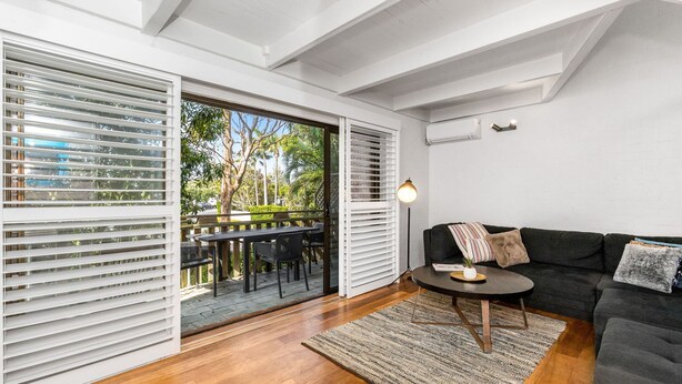A modern living room with a dark grey sofa, a round coffee table, and a large sliding glass door leading to an outdoor area with a wooden deck and greenery. - Byron Bay Accom - Balcony on Lawson - 6 - 34 Lawson Street (Byron Shire)