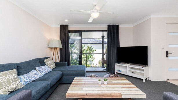 A modern living room with a sectional sofa, a wooden coffee table, a flat-screen TV, and a balcony with curtains. - Byron Bay Accom - Summer Central - Apartment 3  - 5-9 Somerset Street (Byron Shire)