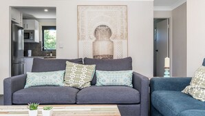 A modern living room with a grey sofa, patterned cushions, a small table with plants, and a kitchen in the background. - Byron Bay Accom - Summer Central - Apartment 3  - 5-9 Somerset Street (Byron Shire)