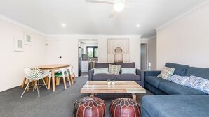 A modern living room with a sofa, coffee table, and ottomans. There is a dining area with a table and chairs. - Byron Bay Accom - Summer Central - Apartment 3  - 5-9 Somerset Street (Byron Shire)