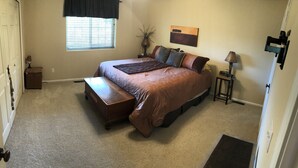 A bedroom with a bed, a bench, a nightstand, a lamp, a window with blinds, and a door. - High Pines Hideaway  on Two Bit Springs Rd. (Deadwood)