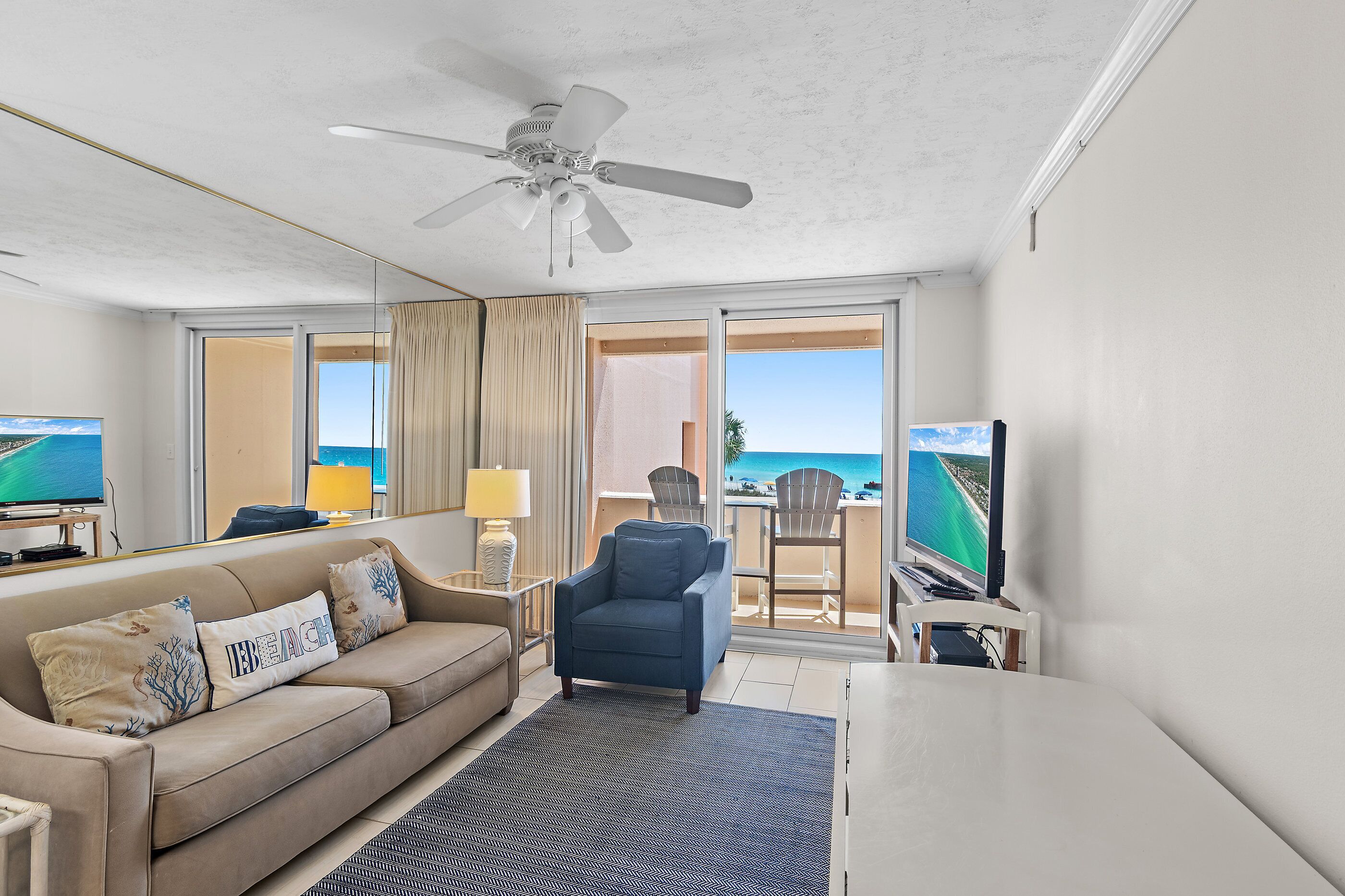 A hotel room with a sofa, armchair, TV, and a balcony with a view of the ocean.