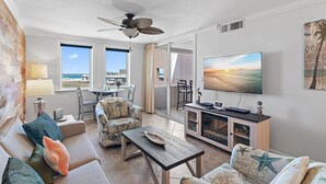 A spacious living room with a large TV, comfortable seating, a ceiling fan, and a view of the ocean through large windows. - Jetty view at Jetty East! One free beach chair set up daily. (Destin)