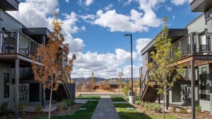 A pathway leading to a modern building with balconies, surrounded by young trees and a clear sky.