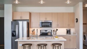 A modern kitchen with a stainless steel refrigerator, a microwave, a stove, and a sink. There are three stools and a table.
