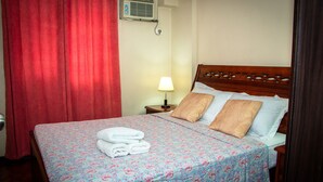 Cama bem arrumada com colcha estampada, duas almofadas e toalhas dobradas em um painel de cama de madeira.