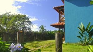 A lush garden with a pathway leading to a building with a blue exterior and a tiled roof. - Recanto dos Dendês Chalés - Chalé Sol - Praia do Sargi, Serra Grande, Uruçuca. (Uruçuca)