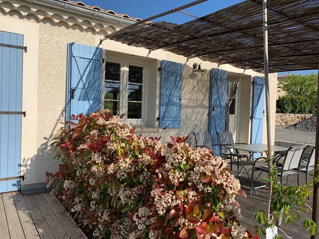 Un espacio exterior techado con una mesa y sillas, una terraza de madera y un edificio con contraventanas azules.
