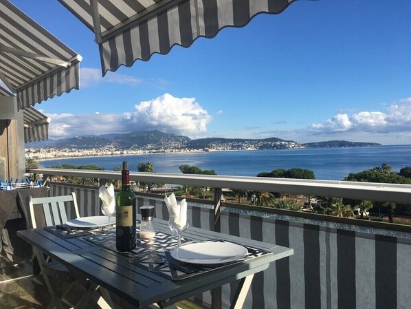 A table set for two with a bottle of wine, glasses, and a napkin on a balcony overlooking a coastal view with mountains in the distance. - 2 Rooms NEW Promenade des Anglais Amazing View (Nice)