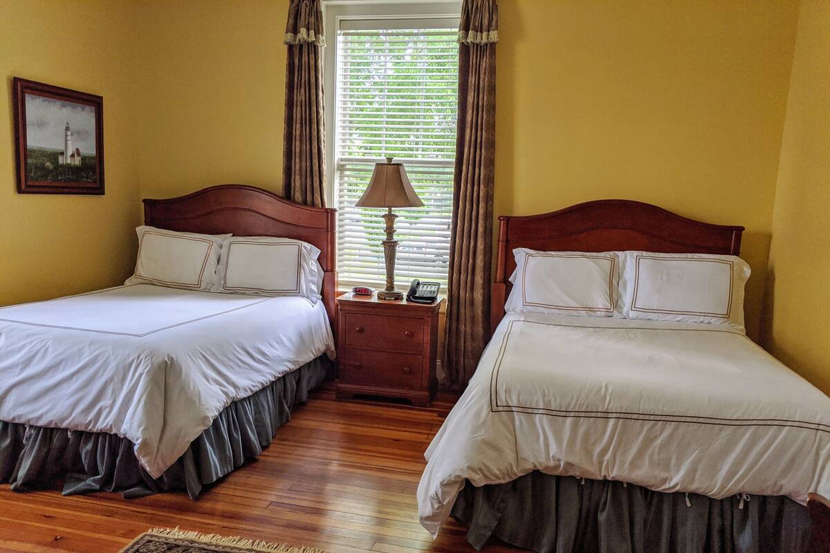 A hotel room with two beds, wooden flooring, a nightstand with a lamp, a framed picture on the wall, and a window with curtains.