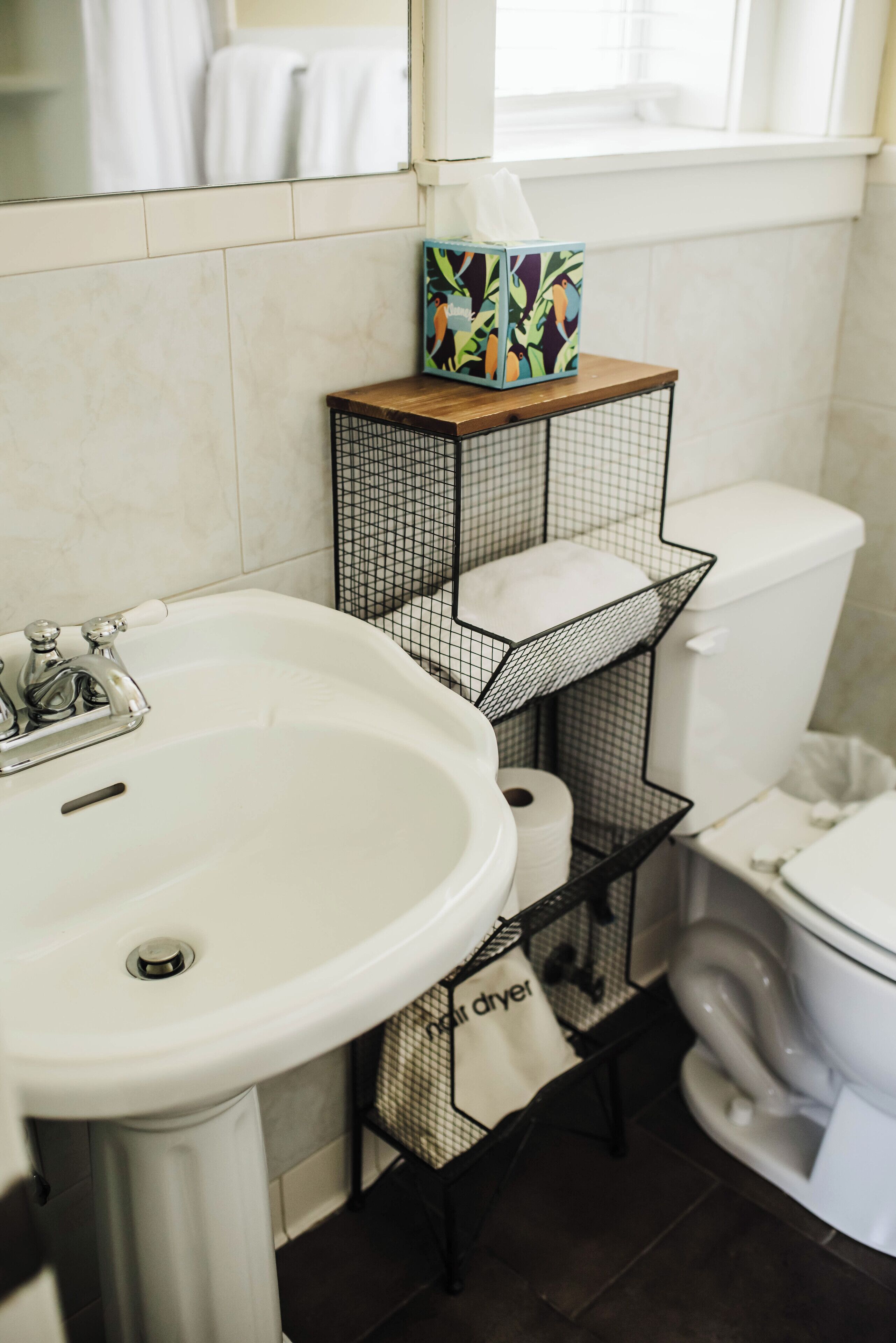Ein Badezimmer mit einem weißen Waschbecken, einer Toilette und einem schwarzen Drahtregal mit Taschentuchbox und weißem Beutel.