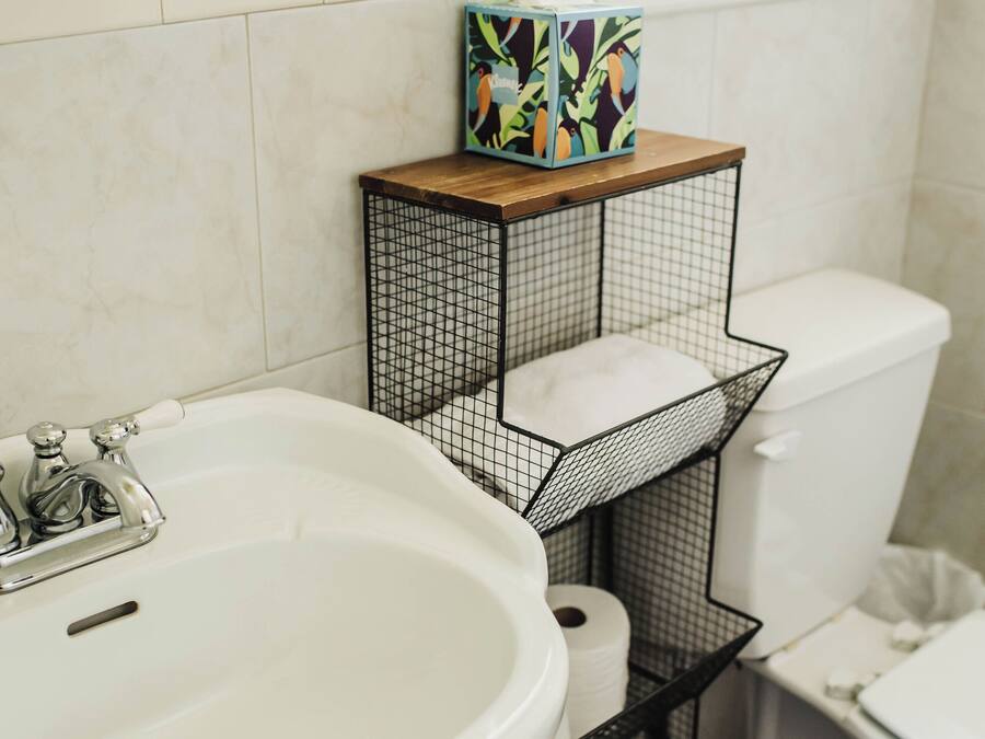 Ein Badezimmer mit einem weißen Waschbecken, einer Toilette und einem schwarzen Drahtregal mit Taschentuchbox und weißem Beutel.