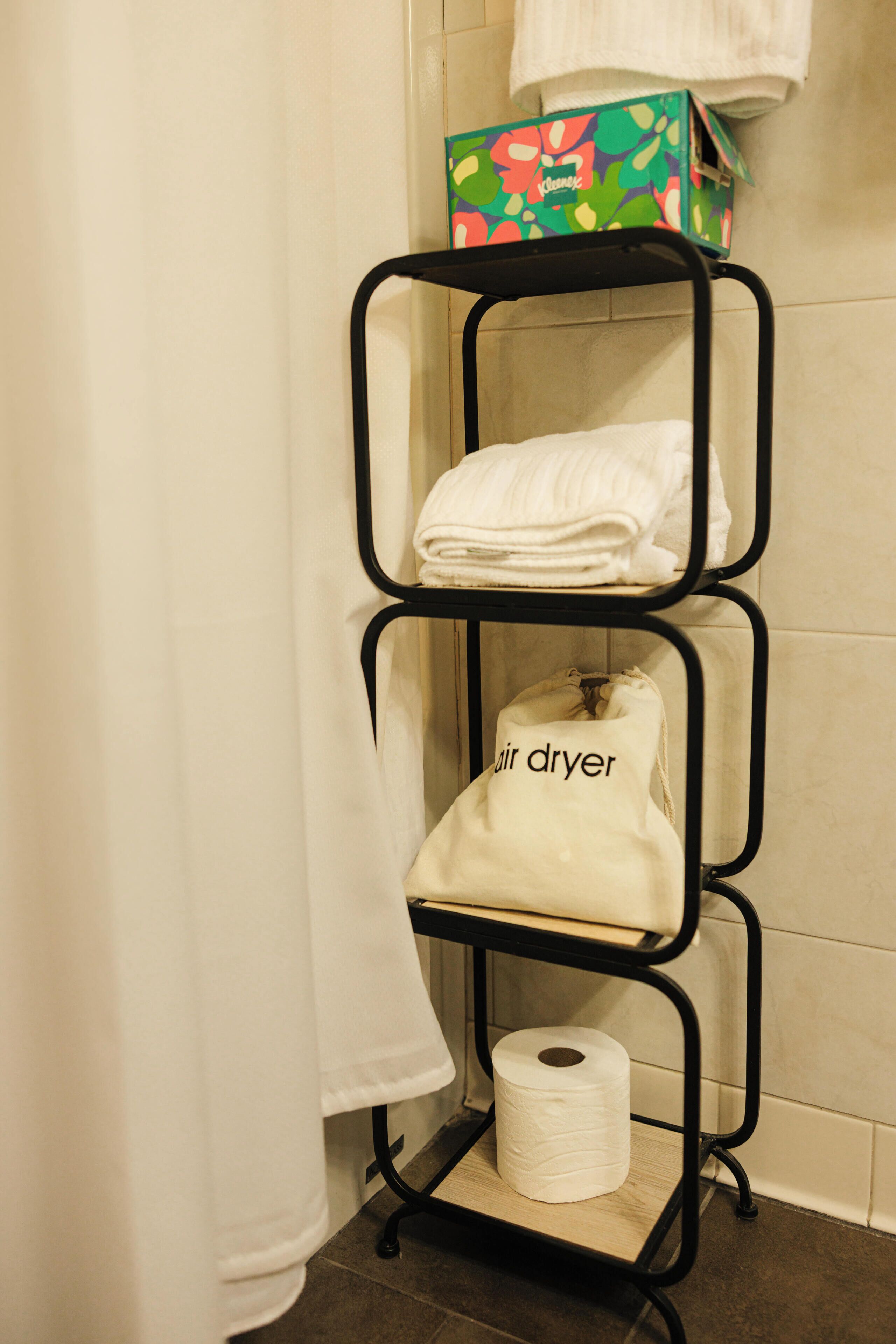 Een badkamer met een metalen plank waarop handdoeken, een pak toiletpapier en een zak met 'air dryer' staan.