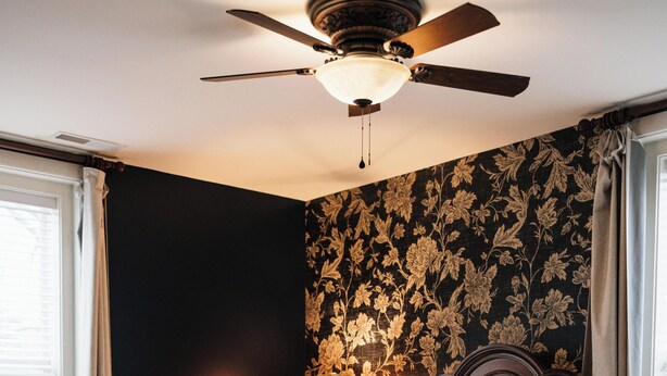 A bedroom with a wooden bed, a ceiling fan, a nightstand, and a wall with floral wallpaper.