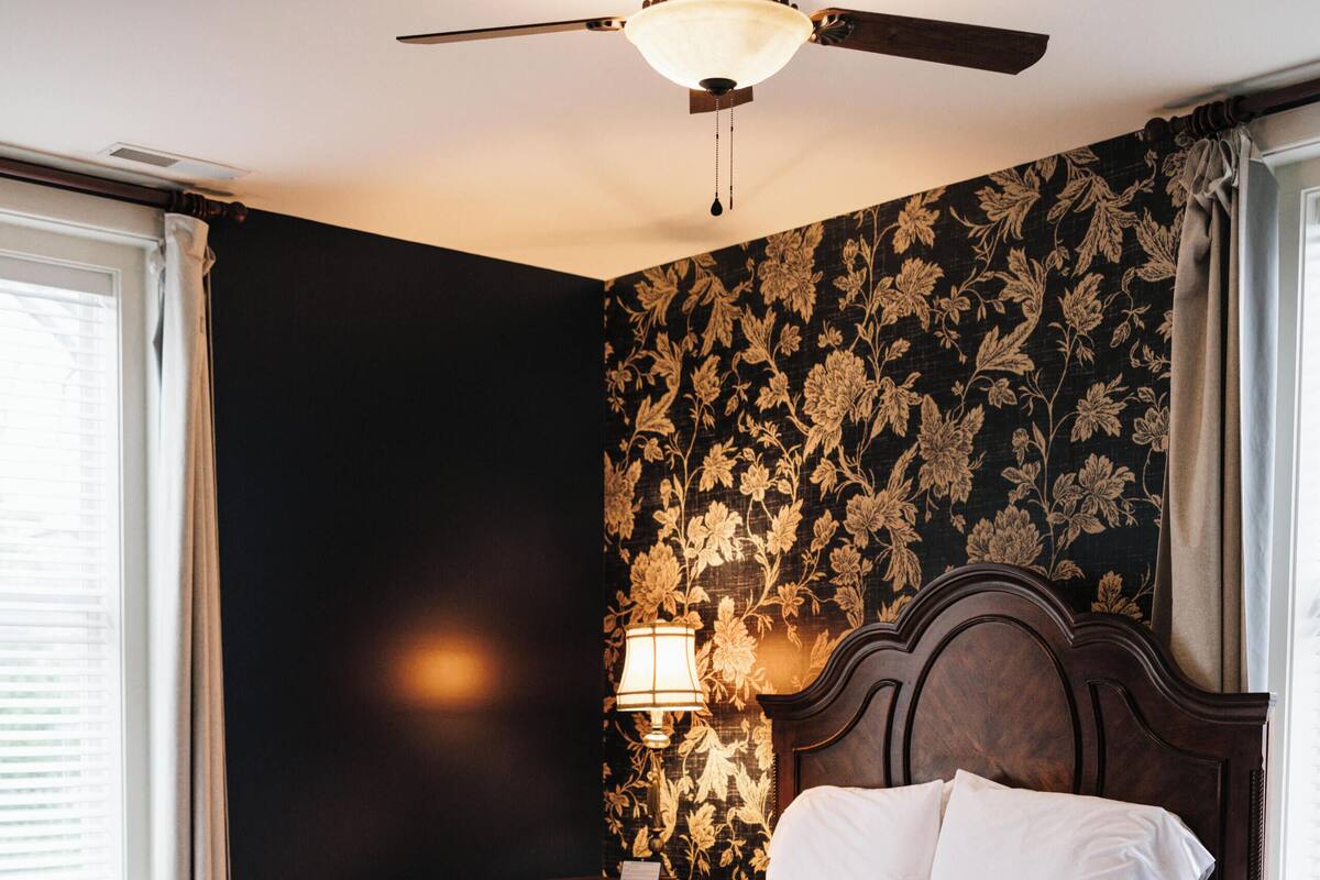 A bedroom with a wooden bed, a ceiling fan, a nightstand, and a wall with floral wallpaper.