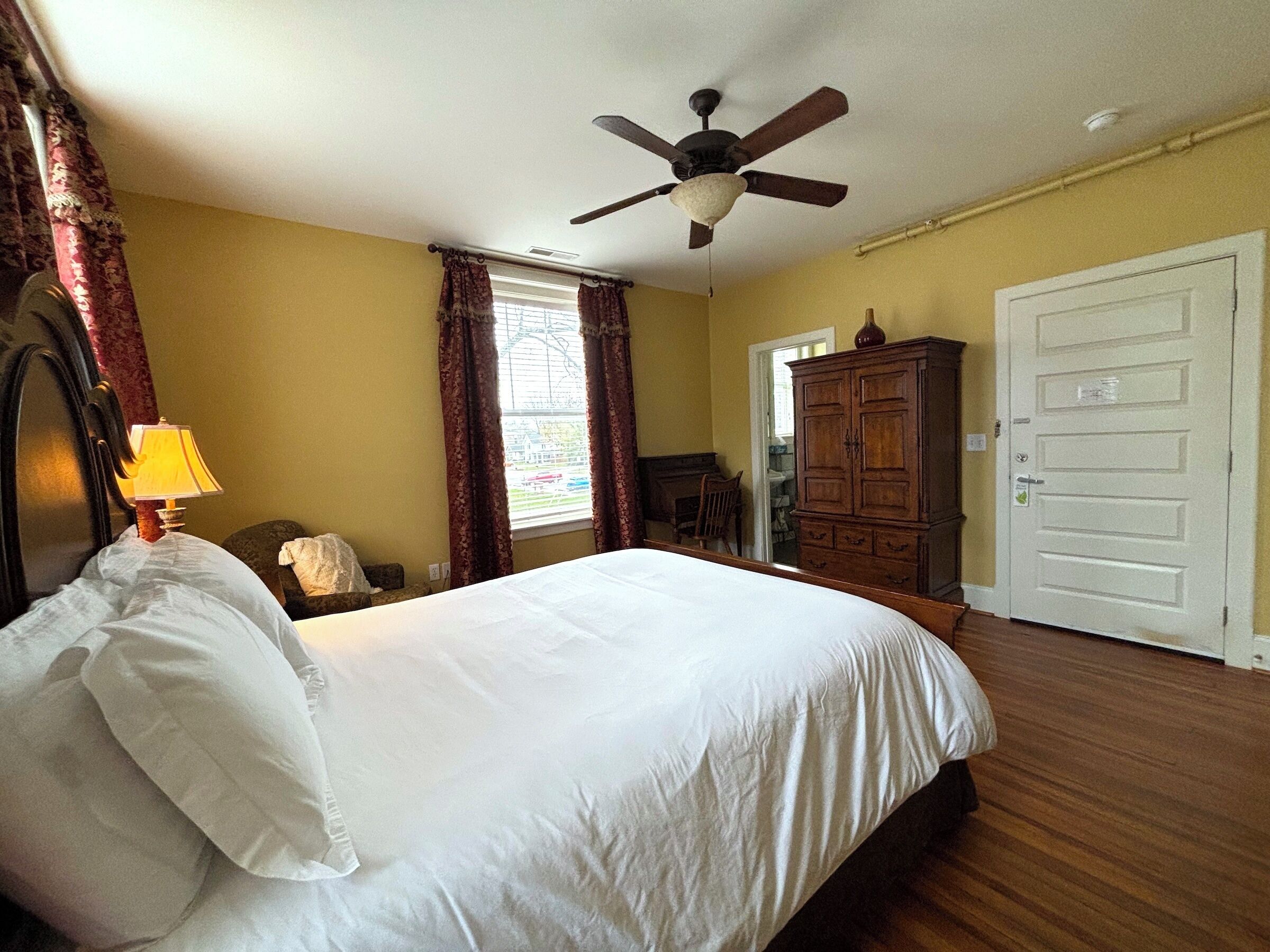 A bedroom with a bed, a ceiling fan, a window with curtains, a wooden wardrobe, and a door.
