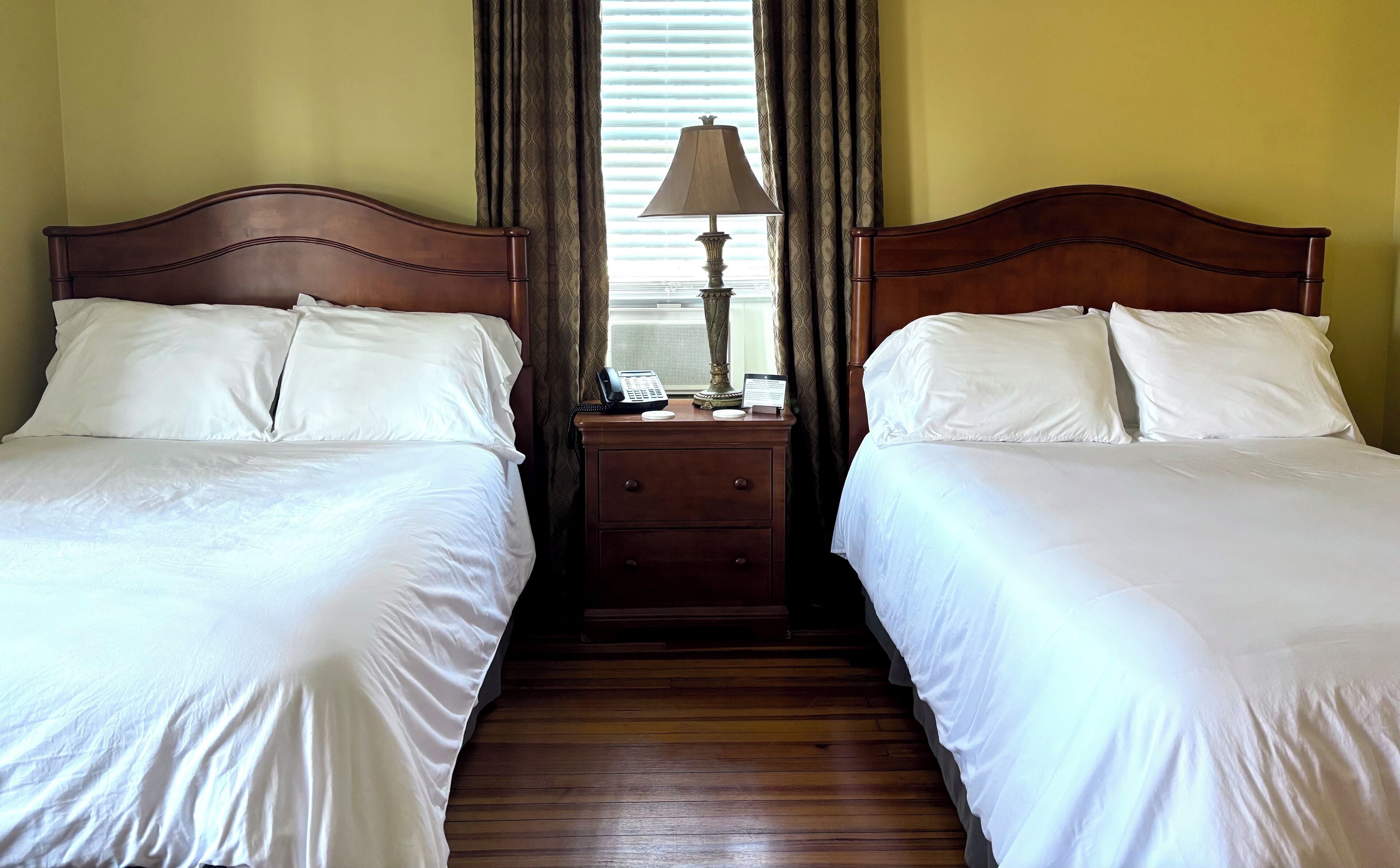 A hotel room with two single beds, a wooden nightstand with a lamp, and a window with blinds.