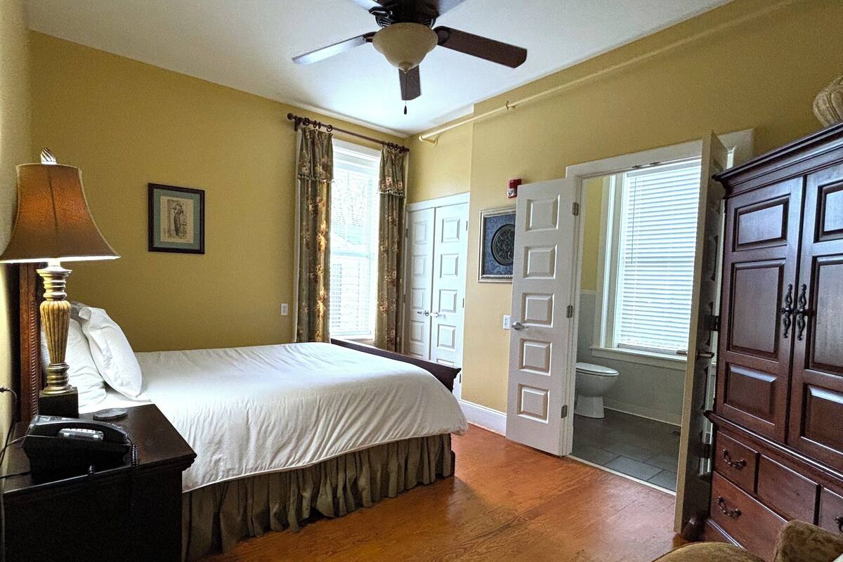 A bedroom with a bed, a bedside lamp, a wooden dresser, and a ceiling fan.