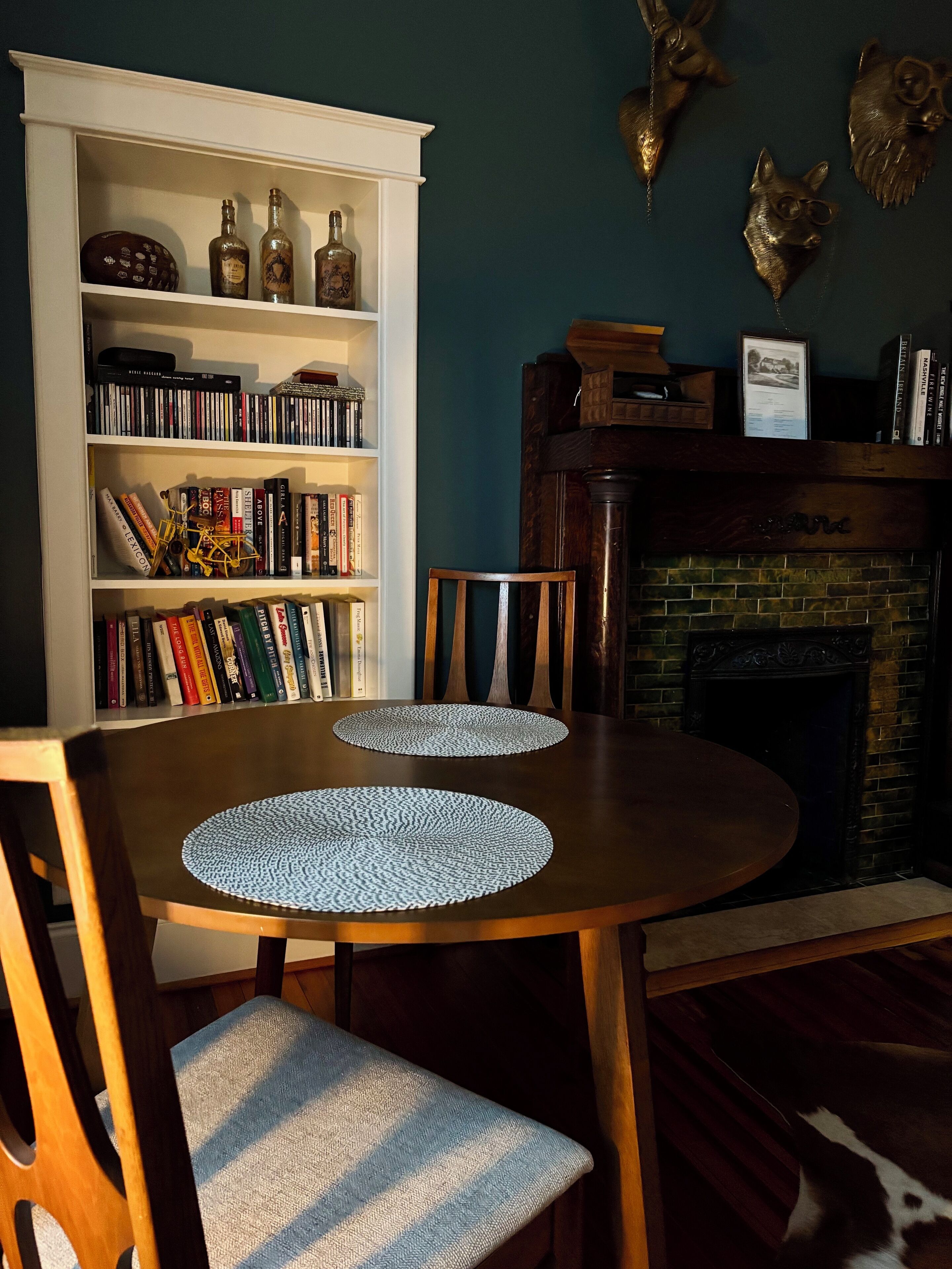 A dining area with a round table, two chairs, a bookshelf with various items, and a fireplace.