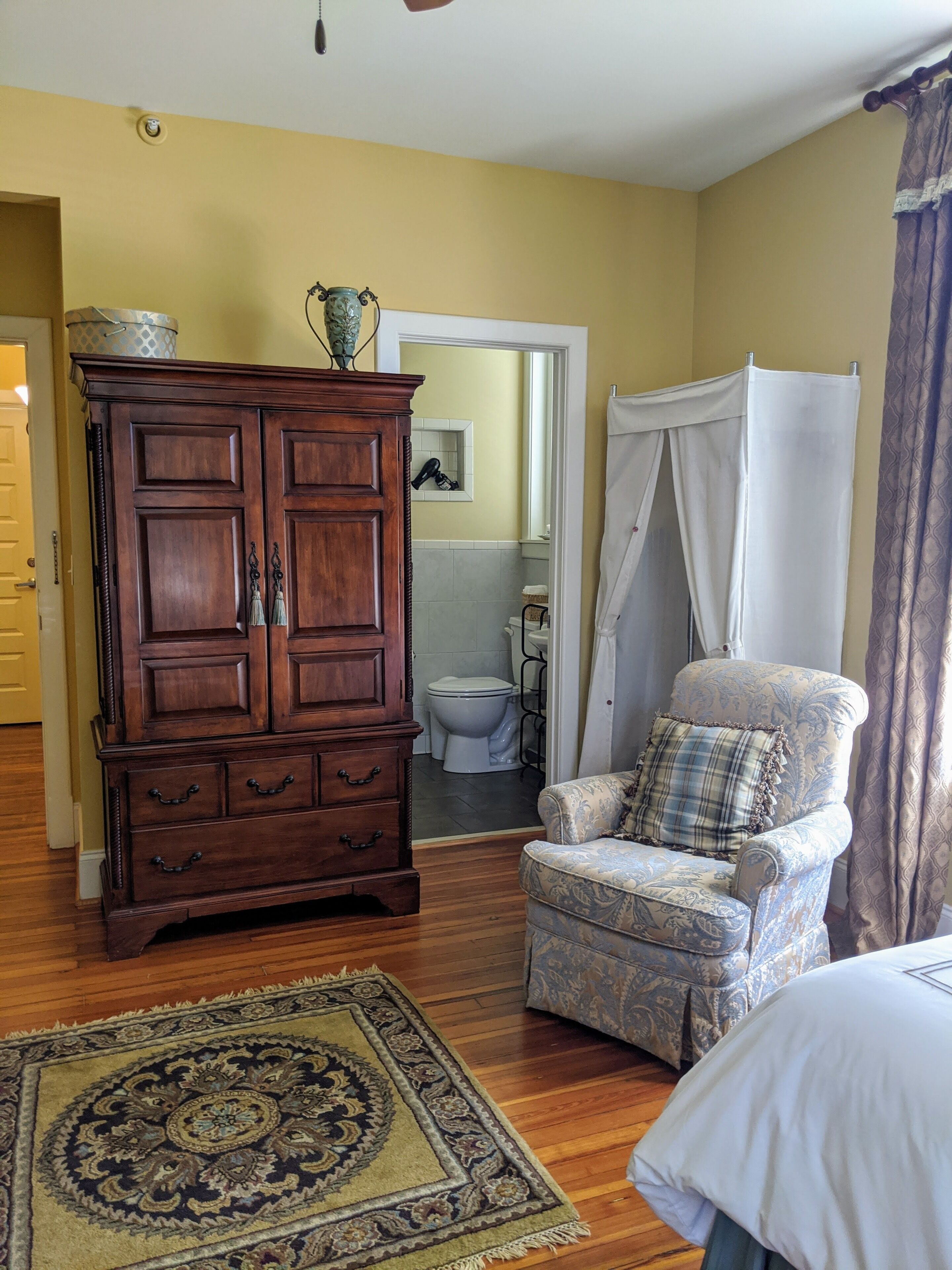 A room with a wooden cabinet, a patterned armchair, a white shower curtain, and a patterned rug.