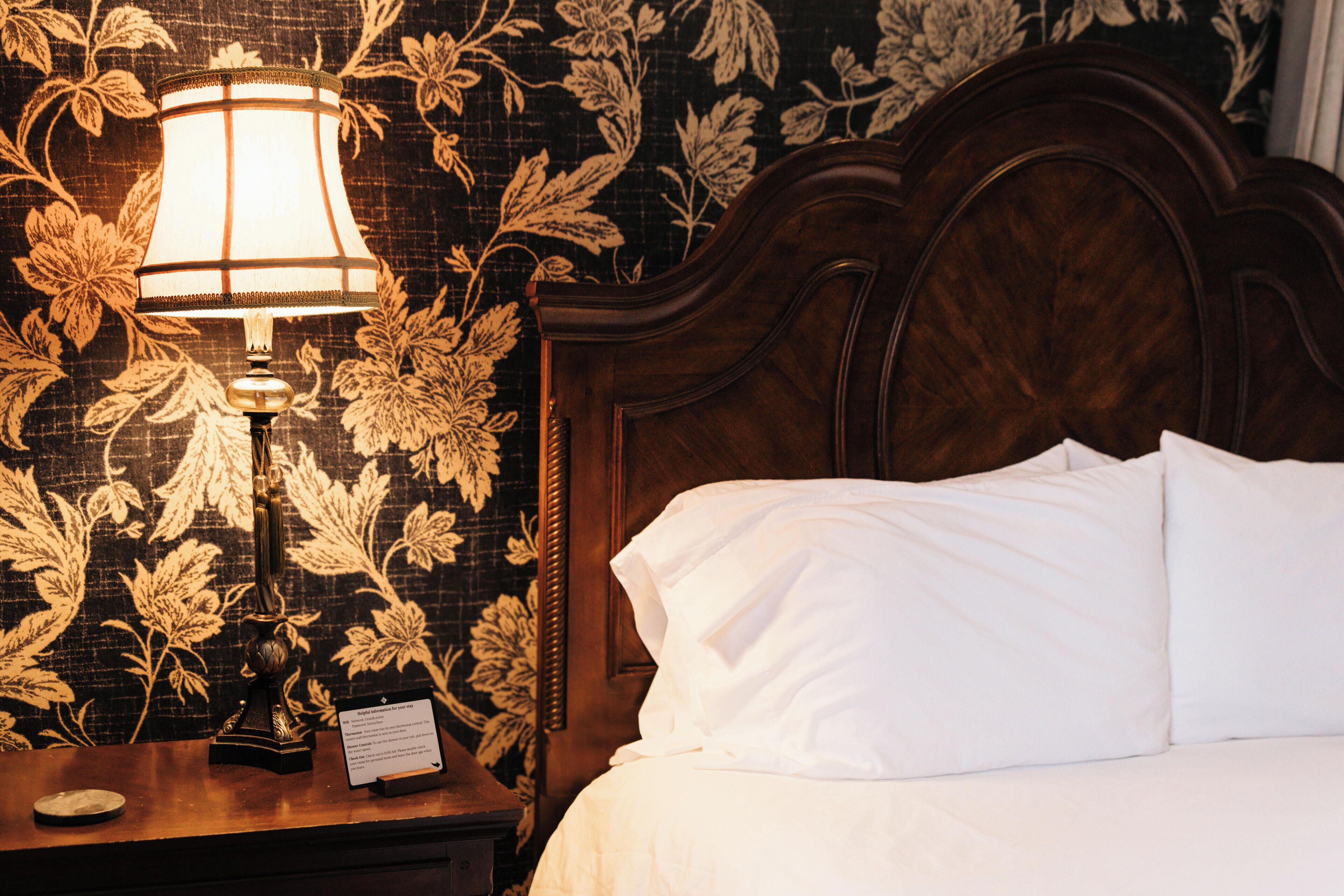 A neatly made bed with white linens, a wooden headboard, and a nightstand with a lamp and a sign.
