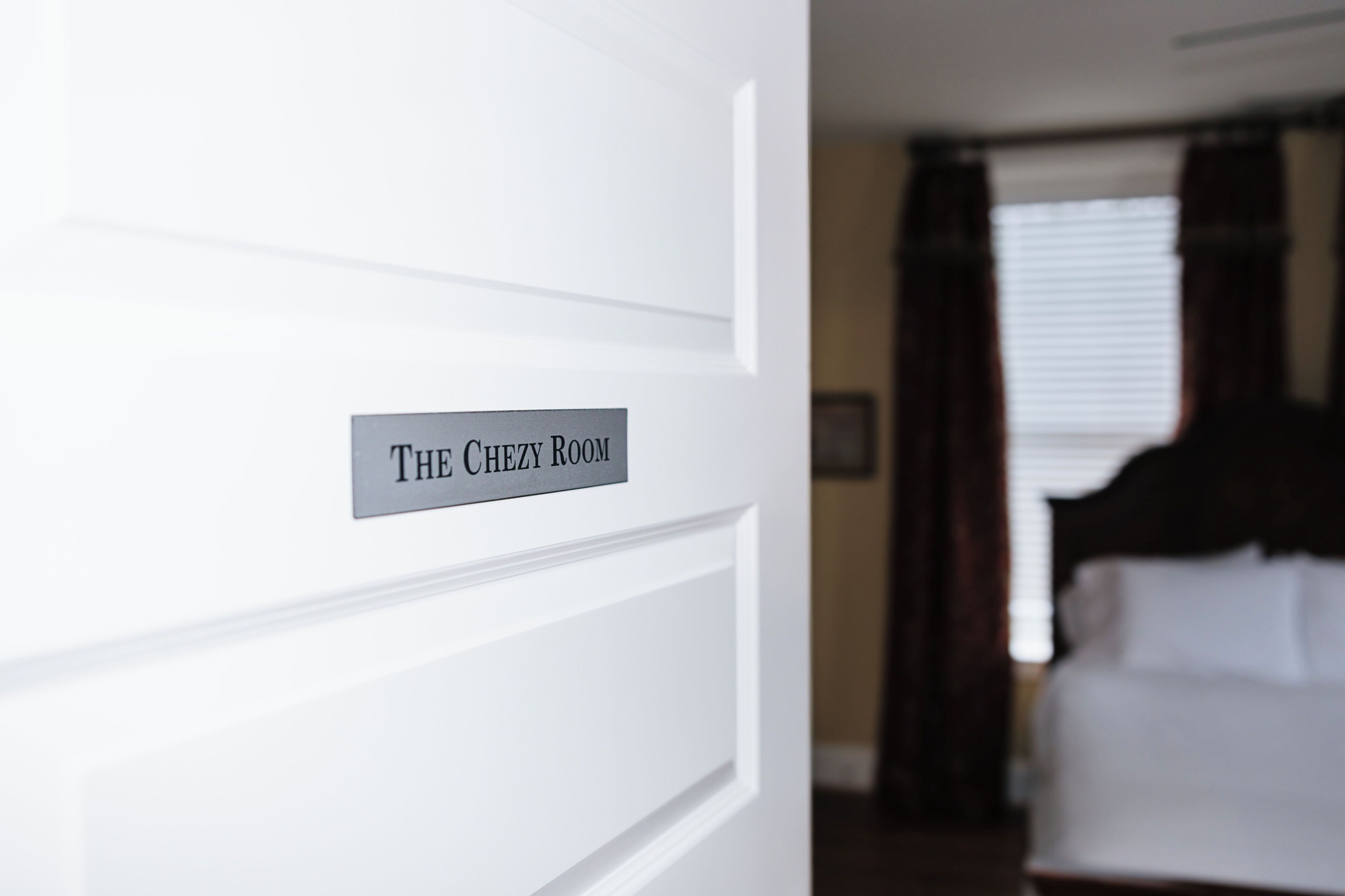 A hotel room with a sign reading 'The Chezy Room,' a bed, and a window with curtains.