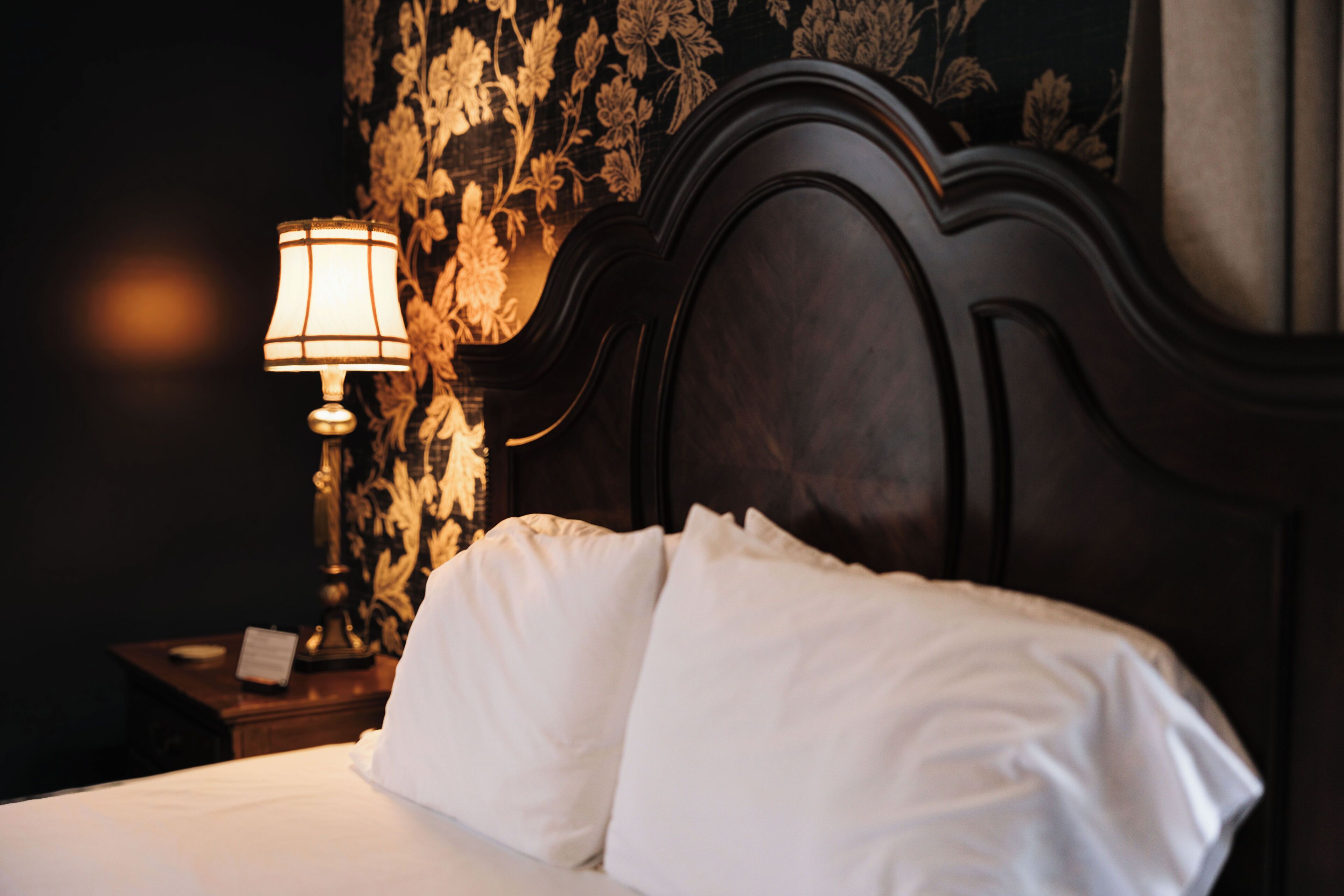 A neatly made bed with white linens, a dark wooden headboard, and a nightstand with a lamp and a small table.
