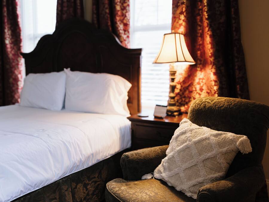 Un letto rifatto con lenzuola bianche, una testiera in legno, un comodino con una lampada e una poltrona confortevole con un cuscino decorativo.
