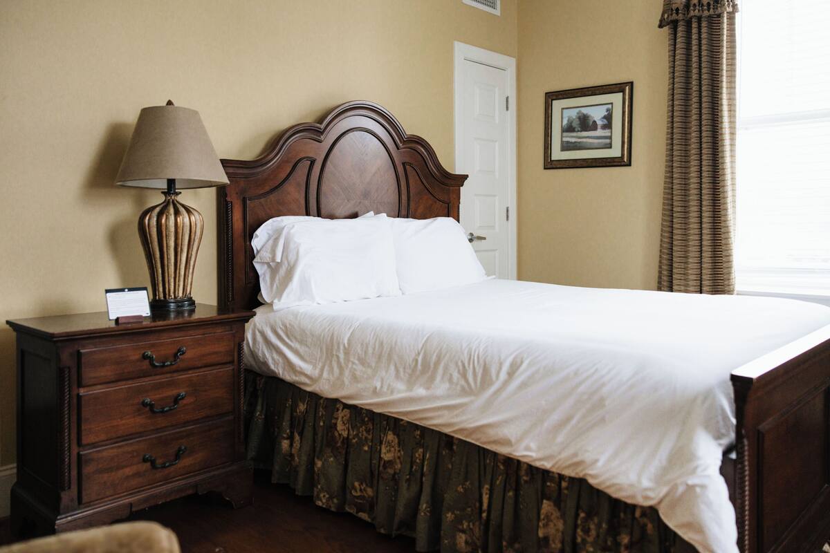 A neatly made bed with a wooden headboard, a bedside table with a lamp, a framed picture on the wall, and a door.