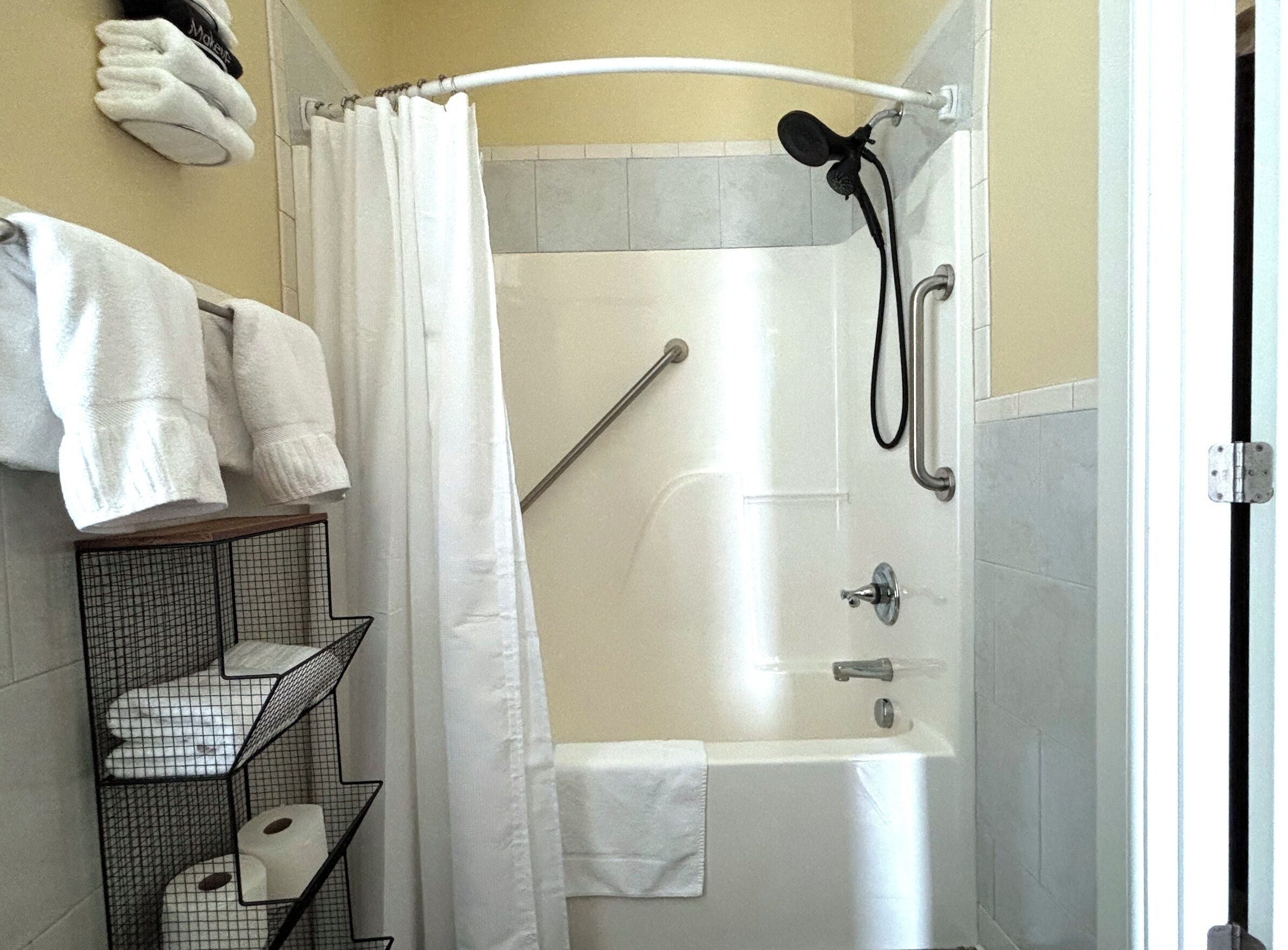 A shower area with a white curtain, a handheld showerhead, and a grab bar. There is a shelf with towels and toilet paper.