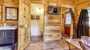 A wooden cabin interior with a bathroom, a sink, a mirror, a television, and a door leading to another room. - Quaint Log Cabin-Fire Pit-Waterside Property-Birds Creek Campground (Sevierville)