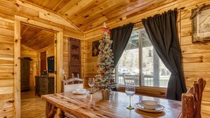 A wooden cabin interior with a dining table set for two, a Christmas tree, and a window with curtains. - Quaint Log Cabin-Fire Pit-Waterside Property-Birds Creek Campground (Sevierville)