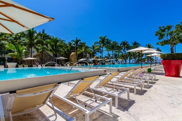 A poolside area with lounge chairs and umbrellas, surrounded by palm trees and a clear blue sky.
