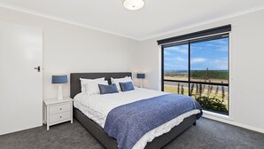 A bedroom with a large window, a bed with white and blue bedding, two bedside tables with lamps, and a view of the outdoors. - Seaclusion - Beachside Luxury (Port Fairy)