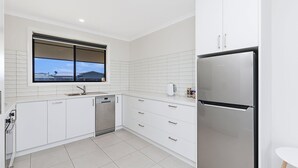 A modern kitchen with white cabinetry, a stainless steel refrigerator, a dishwasher, and a window with a view of buildings. - Seaclusion - Beachside Luxury (Port Fairy)
