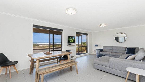 A spacious living area with a large window offering a view of the beach, a grey sofa, a wooden dining table, and a television mounted on the wall. - Seaclusion - Beachside Luxury (Port Fairy)