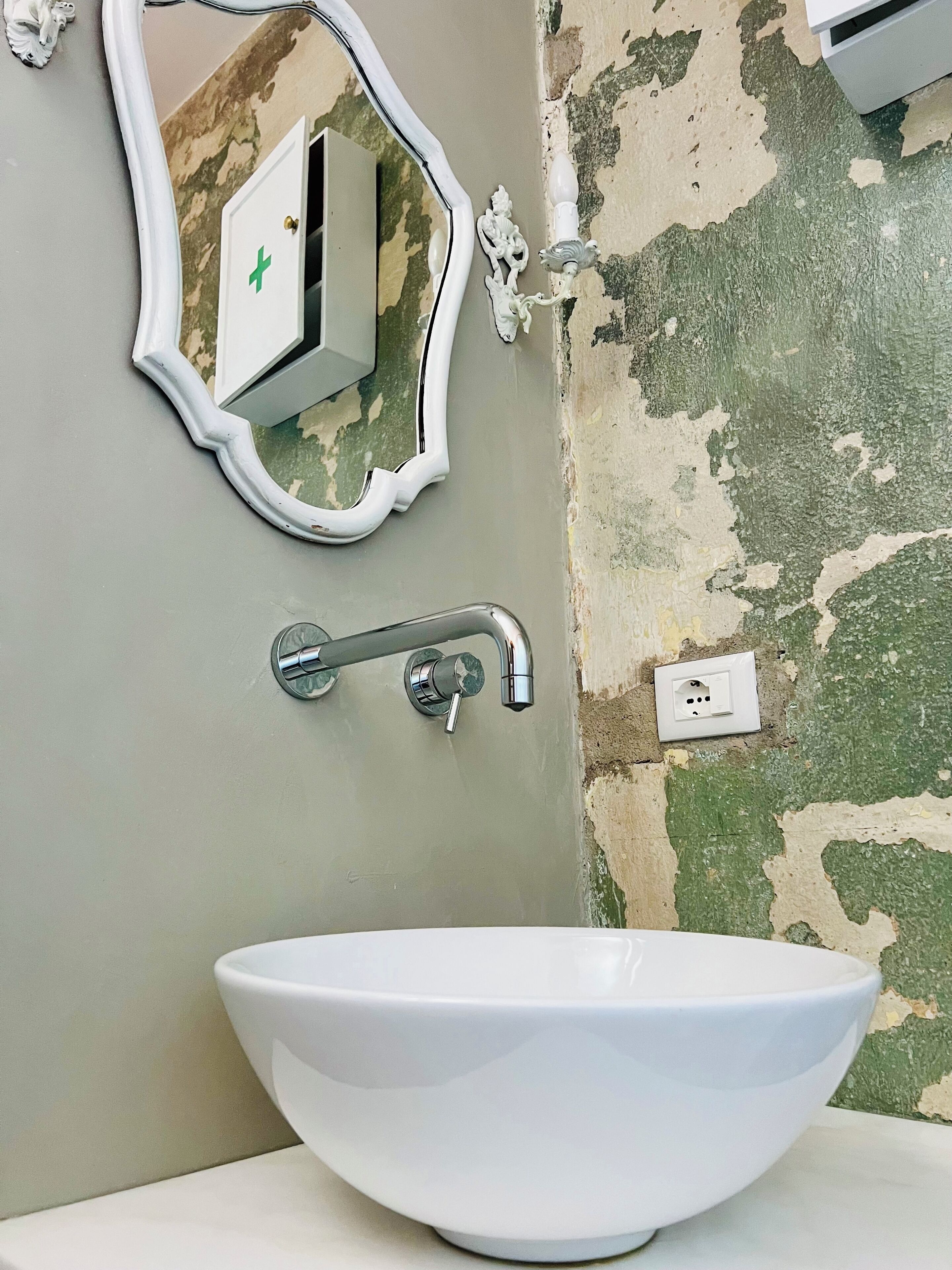 Une salle de bain avec un lavabo blanc, un miroir doté d’un cadre décoratif et un mur dont la peinture s’écaille.