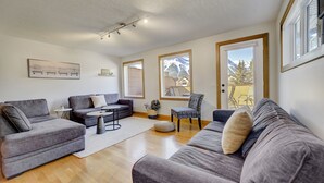 Ein modernes Wohnzimmer mit grauen Sofas, einem Couchtisch und einem gemusterten Sessel. Große Fenster bieten Blick auf die Berge und eine Tür führt nach draußen.