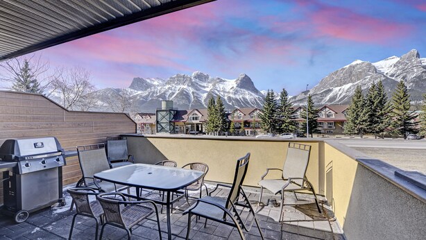 Eine Terrasse im Freien mit Grill, Stühlen und Tisch, mit Blick auf ein Gebirge und eine Stadt mit Gebäuden.