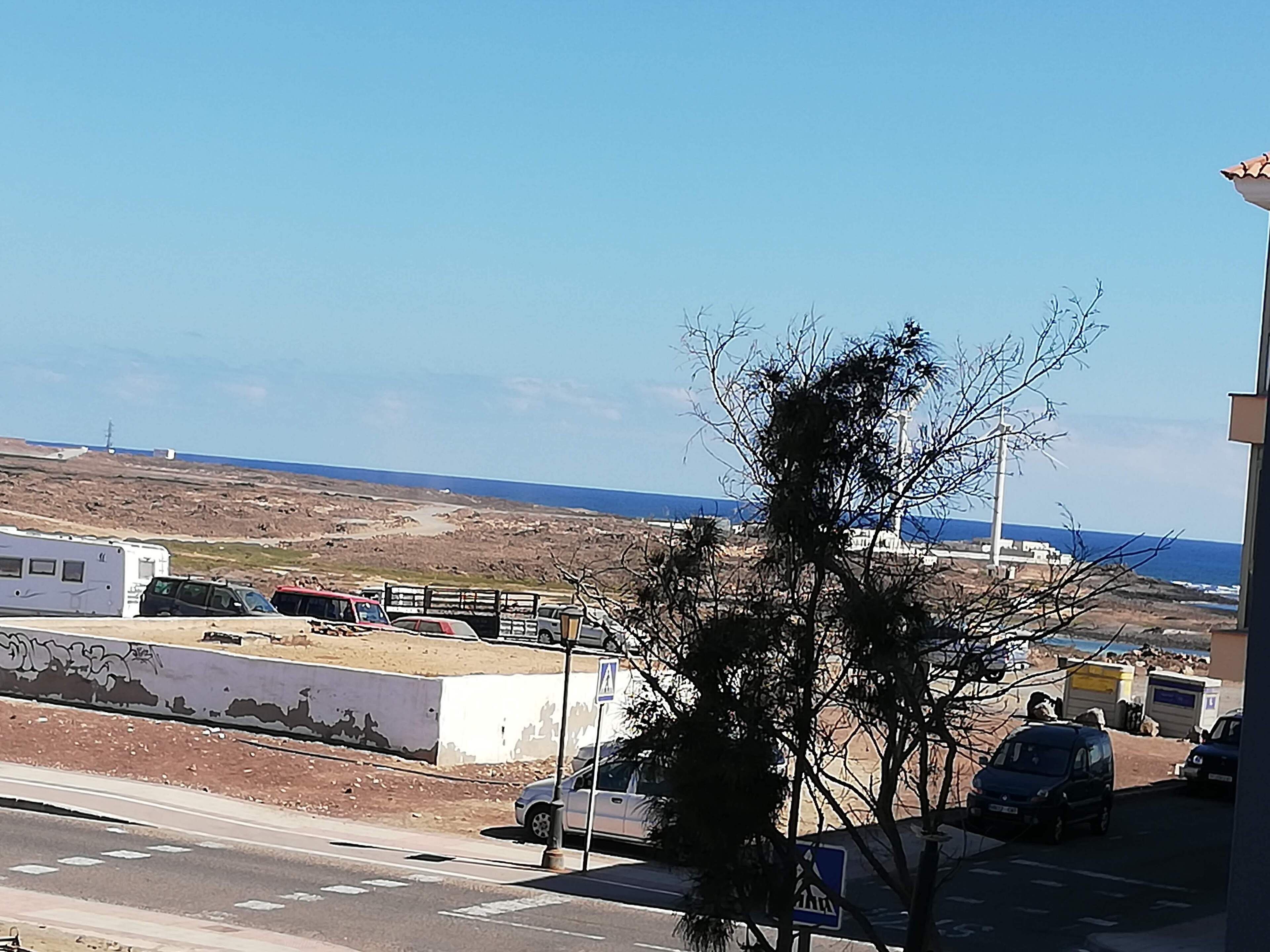 A coastal road with parked vehicles, a pedestrian crossing, and a view of the sea and distant buildings.