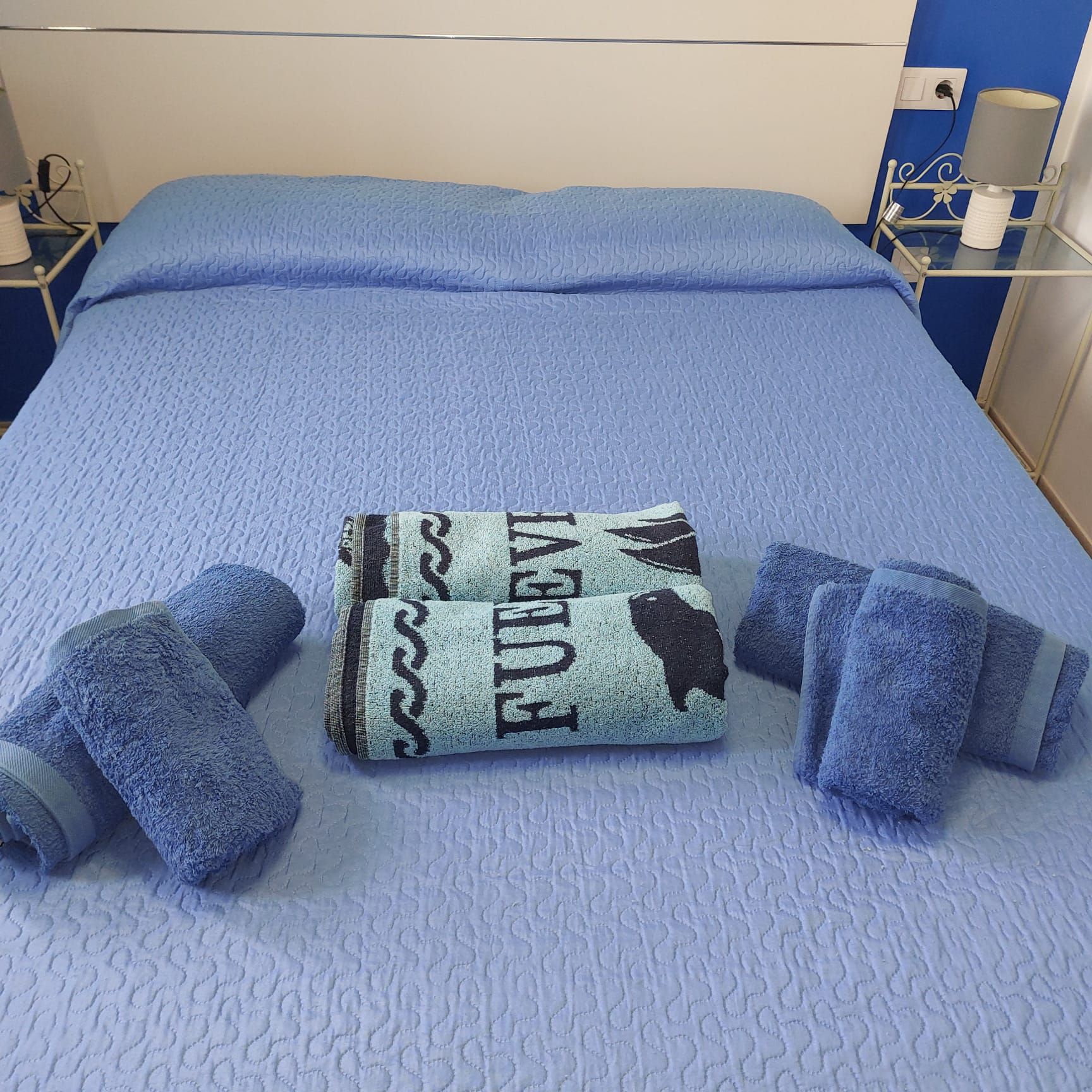 Ein Bett mit einer blauen Steppdecke, ein gemustertes Handtuch gerollt und zwei gefaltete Handtücher.