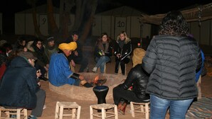 A group of people seated around a campfire in an outdoor setting with numbered tents in the background. - Zagora Desert Camp (Errouha)