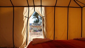 A tent with a bed, a window, and a view of palm trees and a building outside. - Zagora Desert Camp (Errouha)