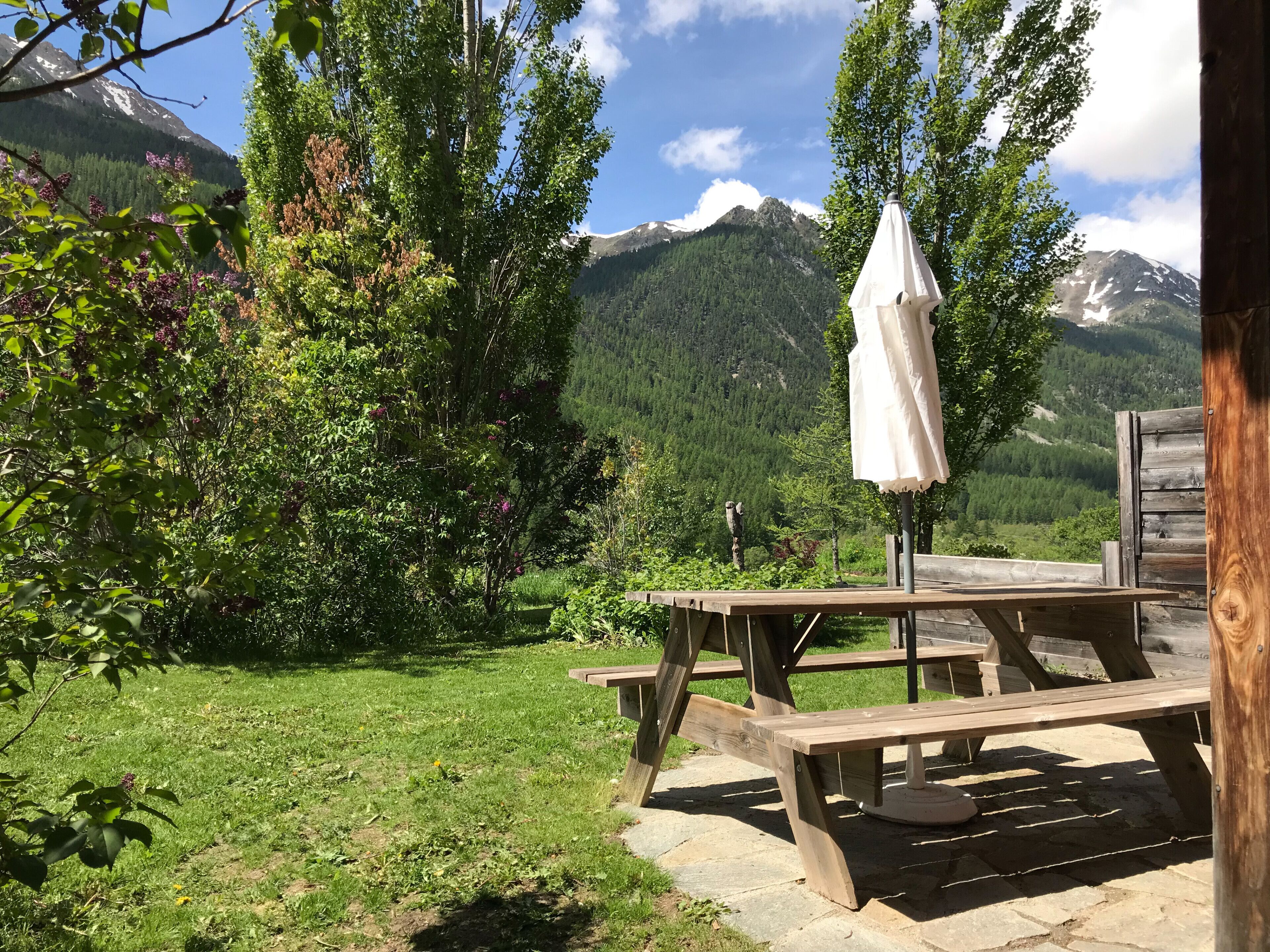 A wooden picnic table with a white umbrella, surrounded by lush greenery and a mountain backdrop.