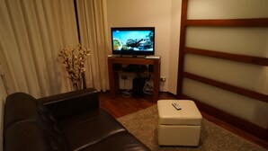 Ein Wohnzimmer mit einem braunen Ledersofa, einem weißen Ottomanen, einem Fernseher auf einem Holzständer und einer Vase mit getrockneten Blumen.